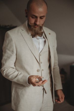 Groom in light beige suit jacket adjusts bow tie while getting ready.