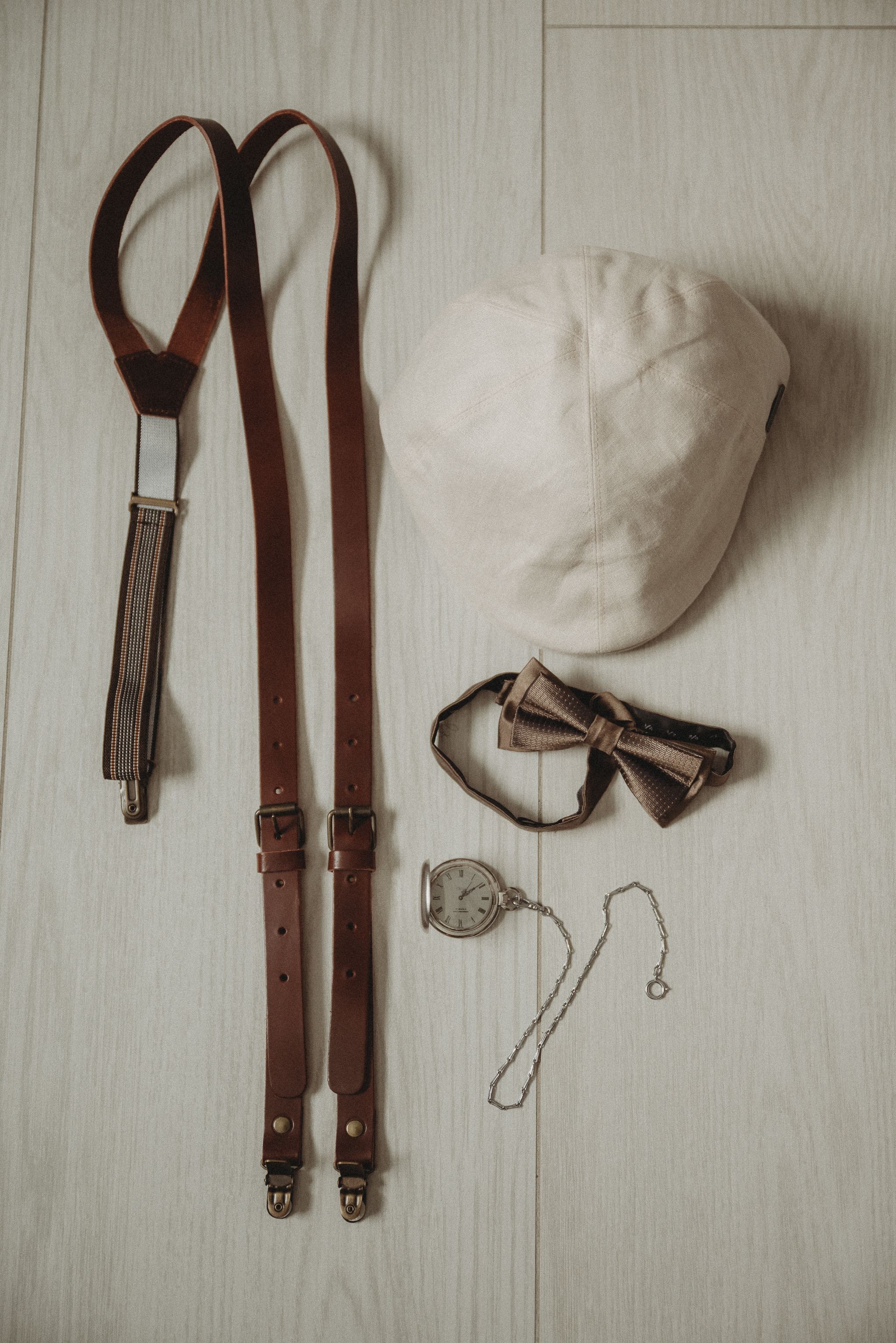 Wedding accessories including suspenders, bow tie, pocket watch and white cap on wooden background.