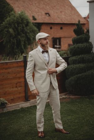 Vintage-styled groom in light suit and flat cap poses in garden with brick building backdrop.