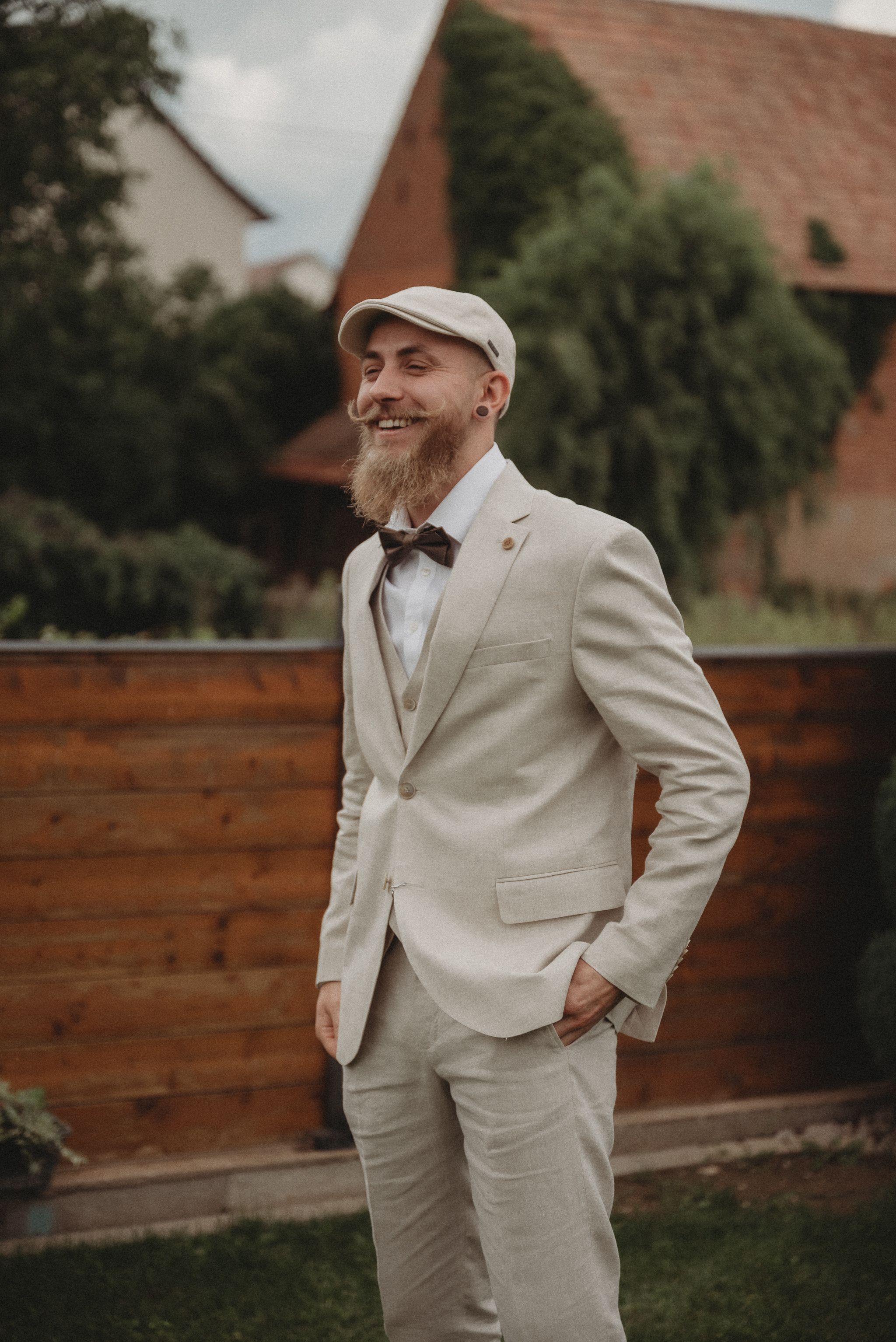 Groom in beige suit and flat cap smiles joyfully while posing outdoors against wooden fence.
