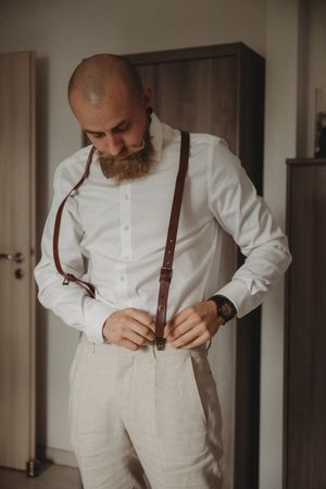 A stylish groom adjusts brown leather suspenders over a crisp white shirt and light-colored trousers.