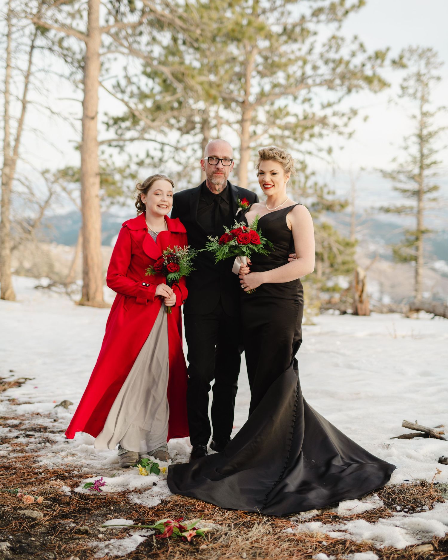 Three people in formal attire pose for a winter wedding photo with snow and pine trees in the background.