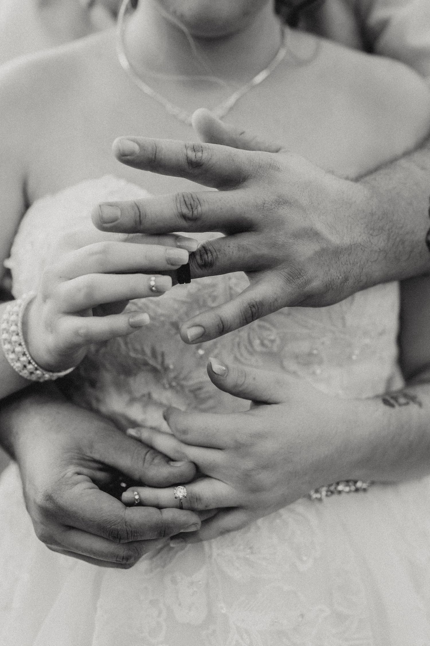 Close-up black and white shot of hands wearing wedding rings and embracing against a white dress.