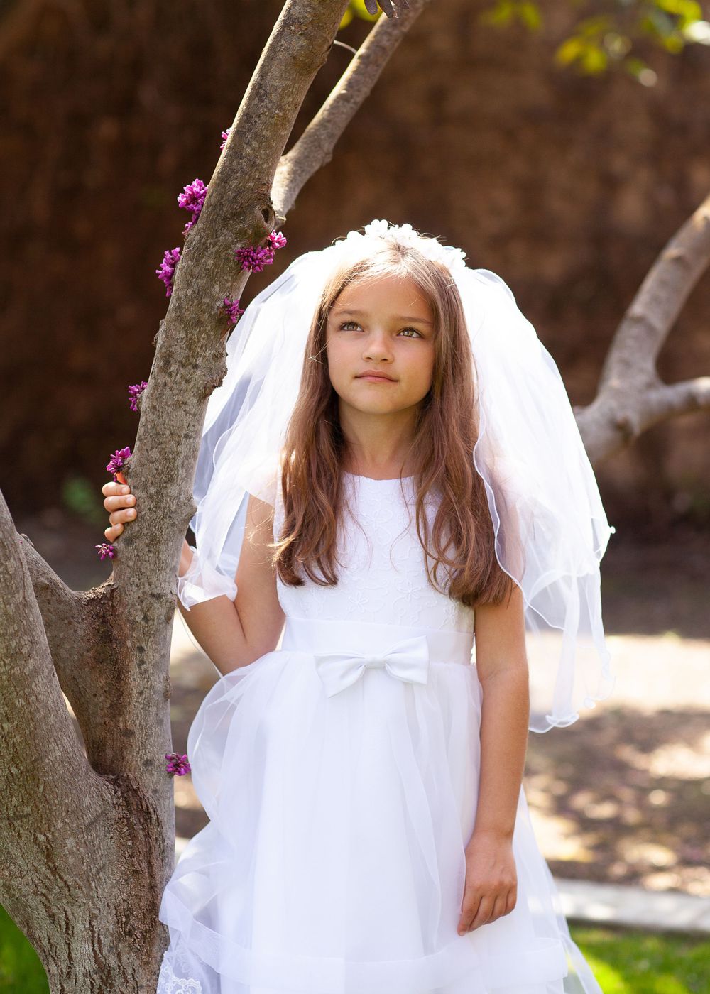 Young person in white communion or flower girl dress poses by flowering tree.
