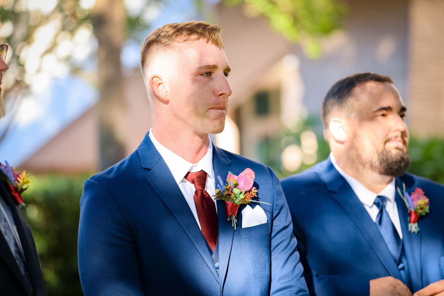Groomsmen in navy suits with red accents stand during outdoor wedding ceremony.