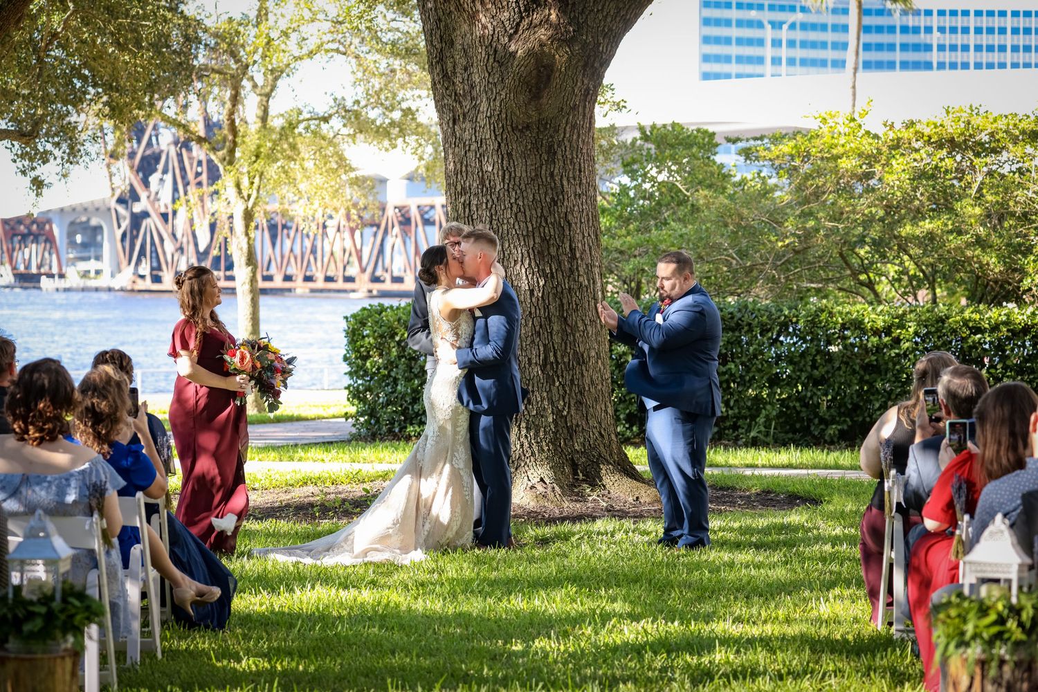 An outdoor wedding ceremony taking place under a large tree by the waterfront.