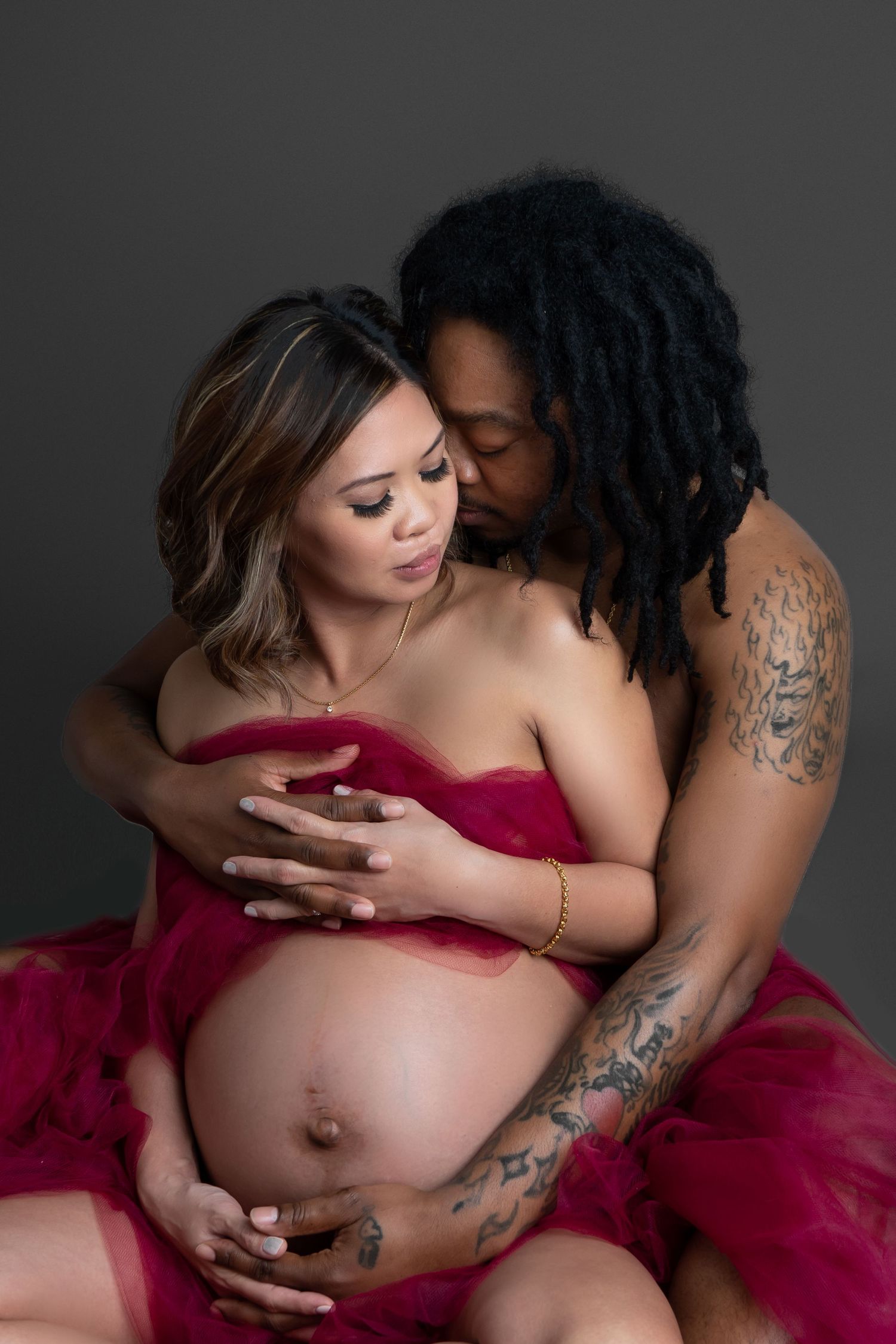 Intimate maternity photoshoot featuring couple in red tulle dress against dark backdrop.