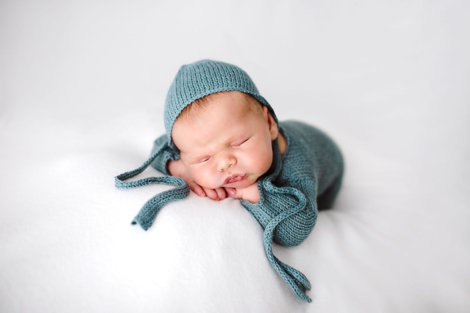 Adorable sleeping newborn baby wearing a teal knit outfit and bonnet on a white background.