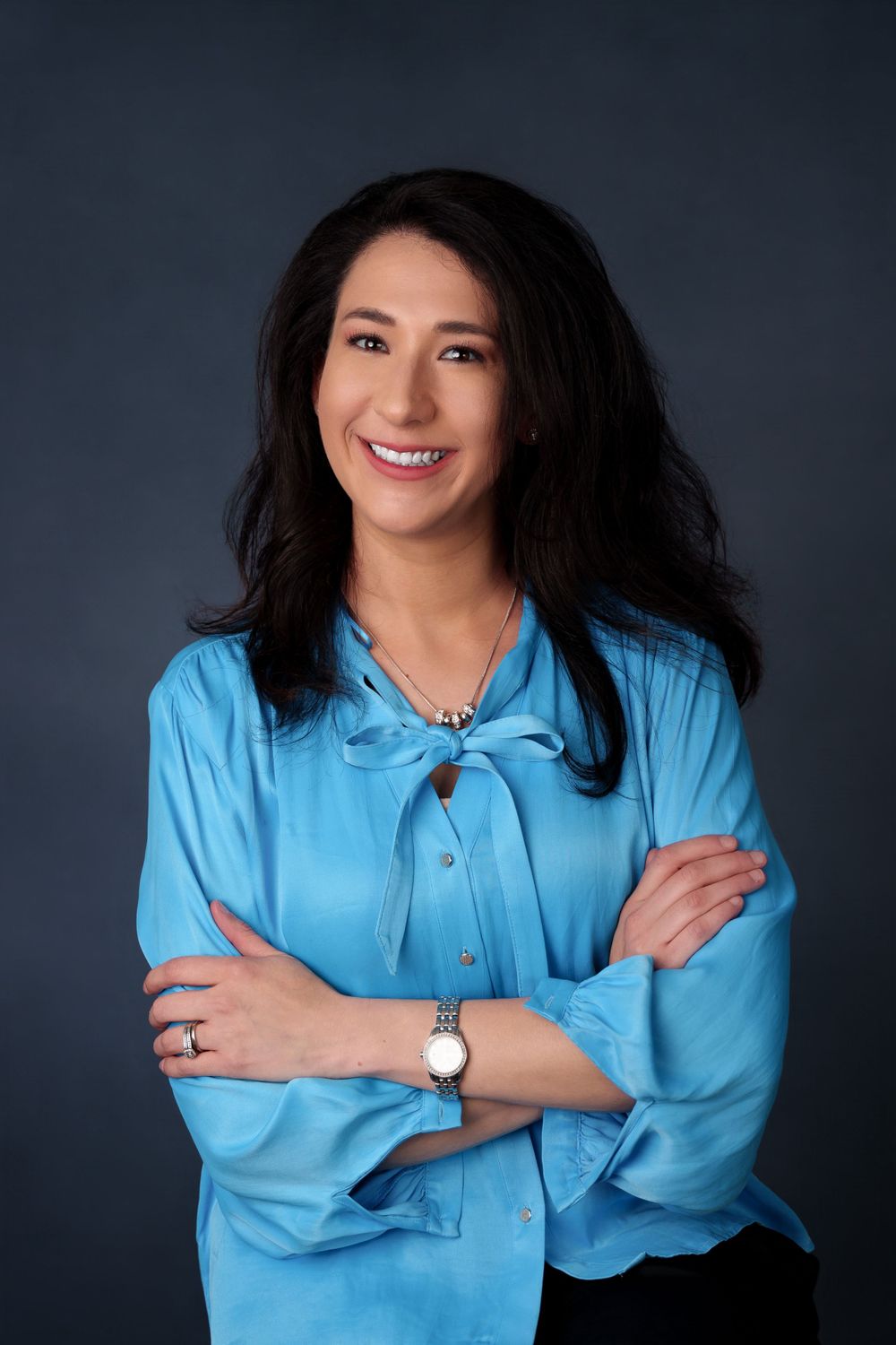 Professional portrait against dark background featuring bright blue blouse and elegant jewelry.