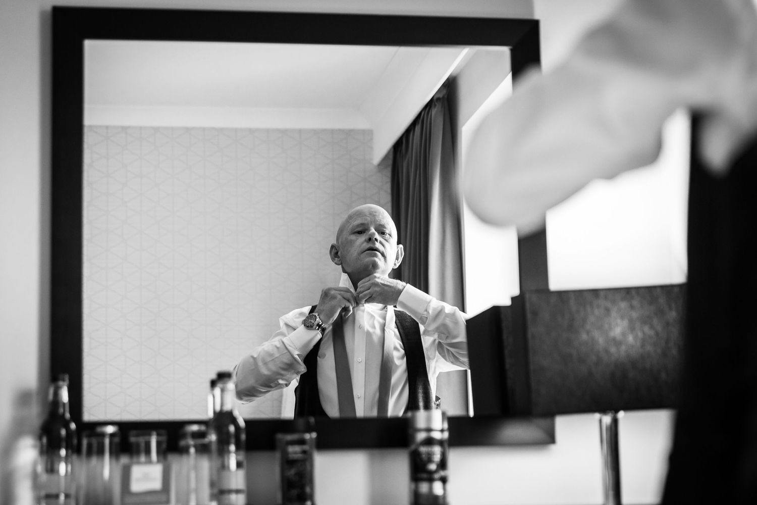 Groom stands looking in the mirror fastening his tie on the wedding morning at Liverpool Crowne Plaza hotel.