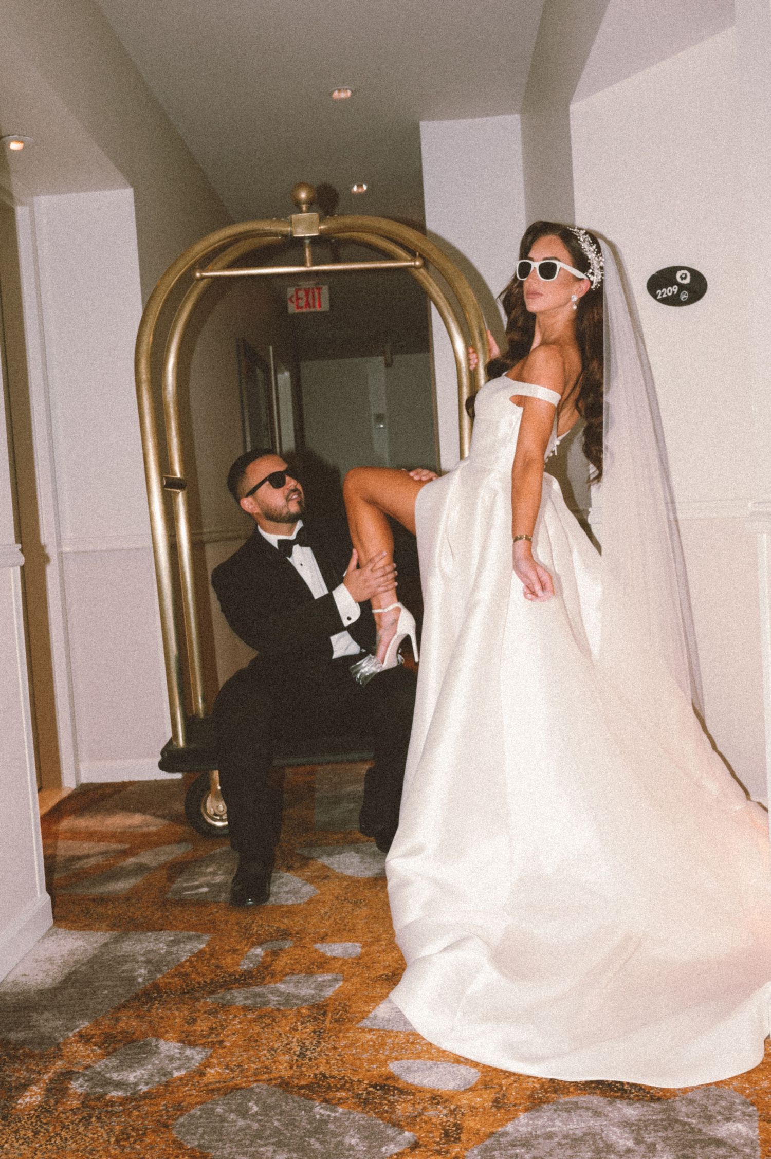 Playful wedding photo under gold archway with bride in sunglasses and off-shoulder gown.