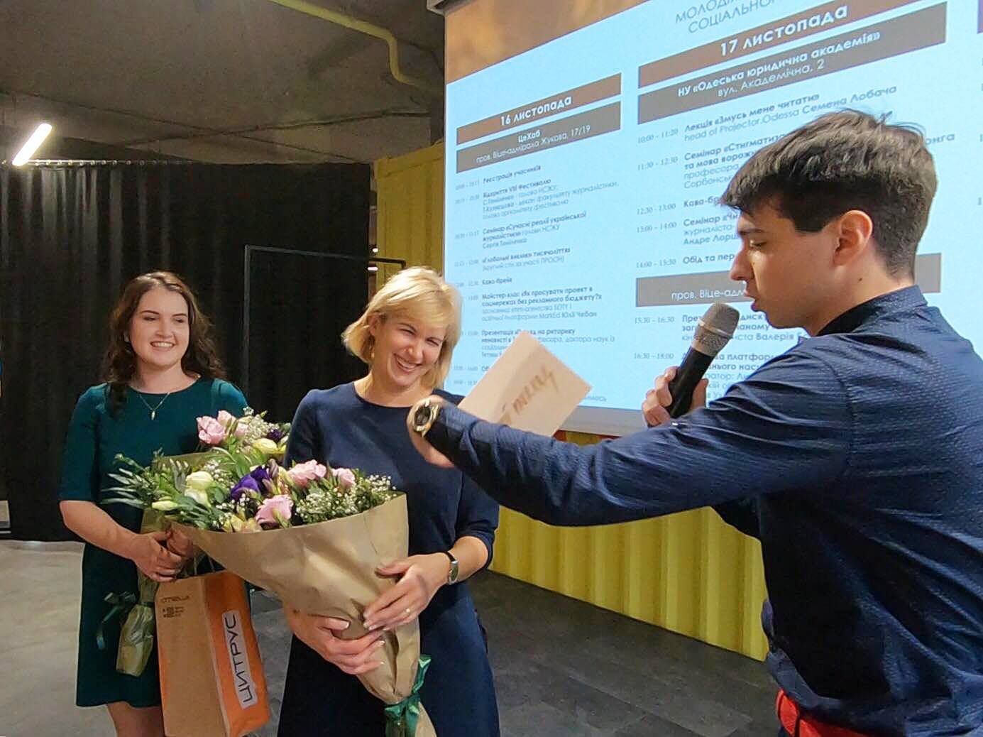 Presentation event with a bouquet being handed over in front of a projected slide.