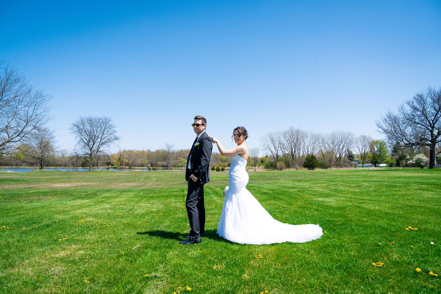 Newlyweds pose together on green lawn under bright blue sky.
