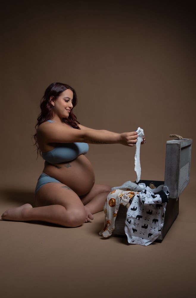 Maternity photo of expecting mother sorting baby clothes against brown backdrop.