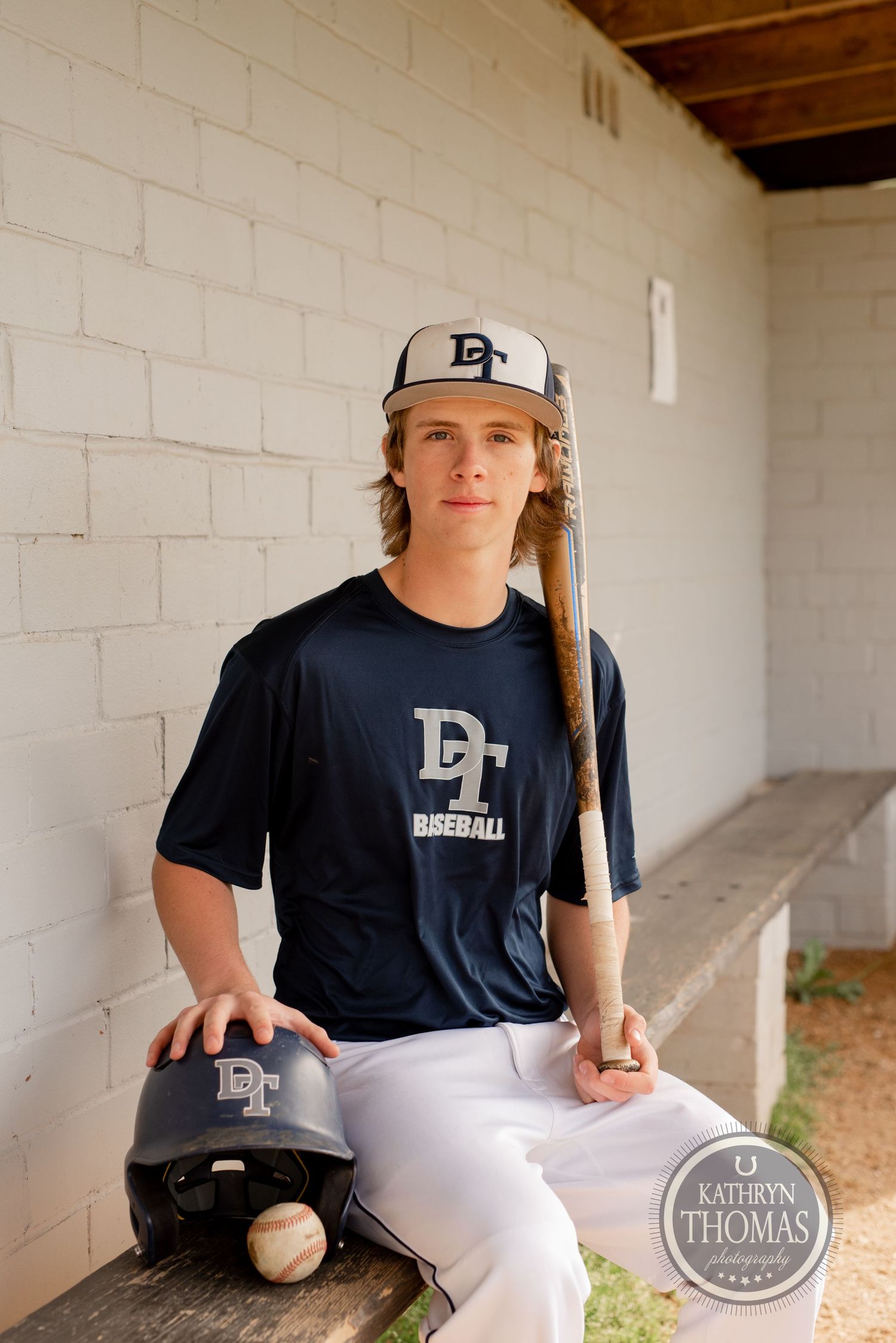 Baseball player sits in dugout with bat and helmet wearing navy team uniform.