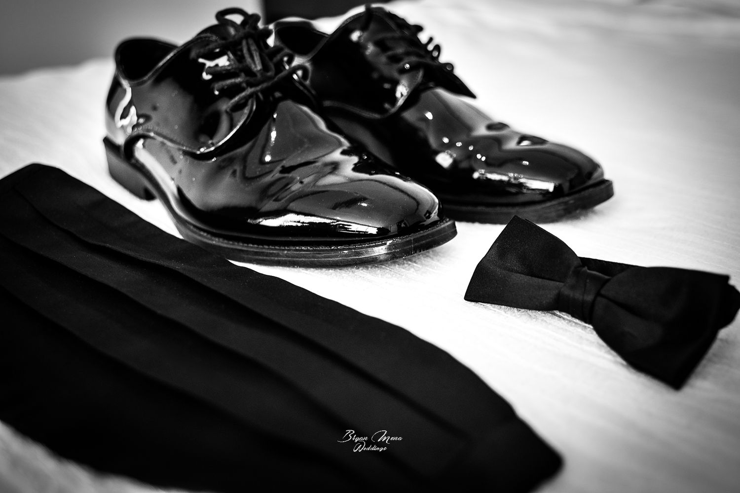 Formal black patent leather dress shoes and bow tie arranged on white surface.