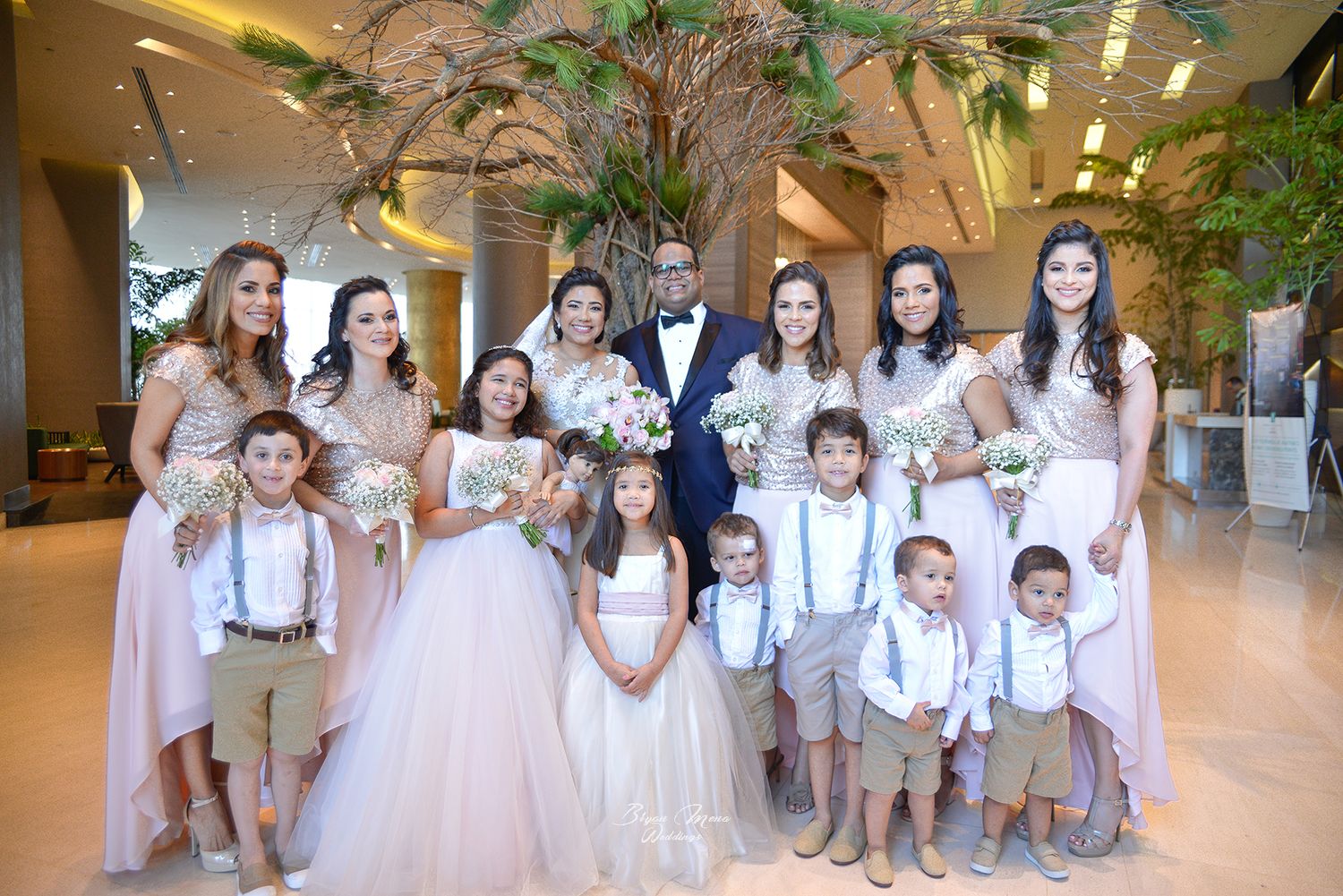 Wedding party in pink dresses, white gowns, and suspenders poses indoors.
