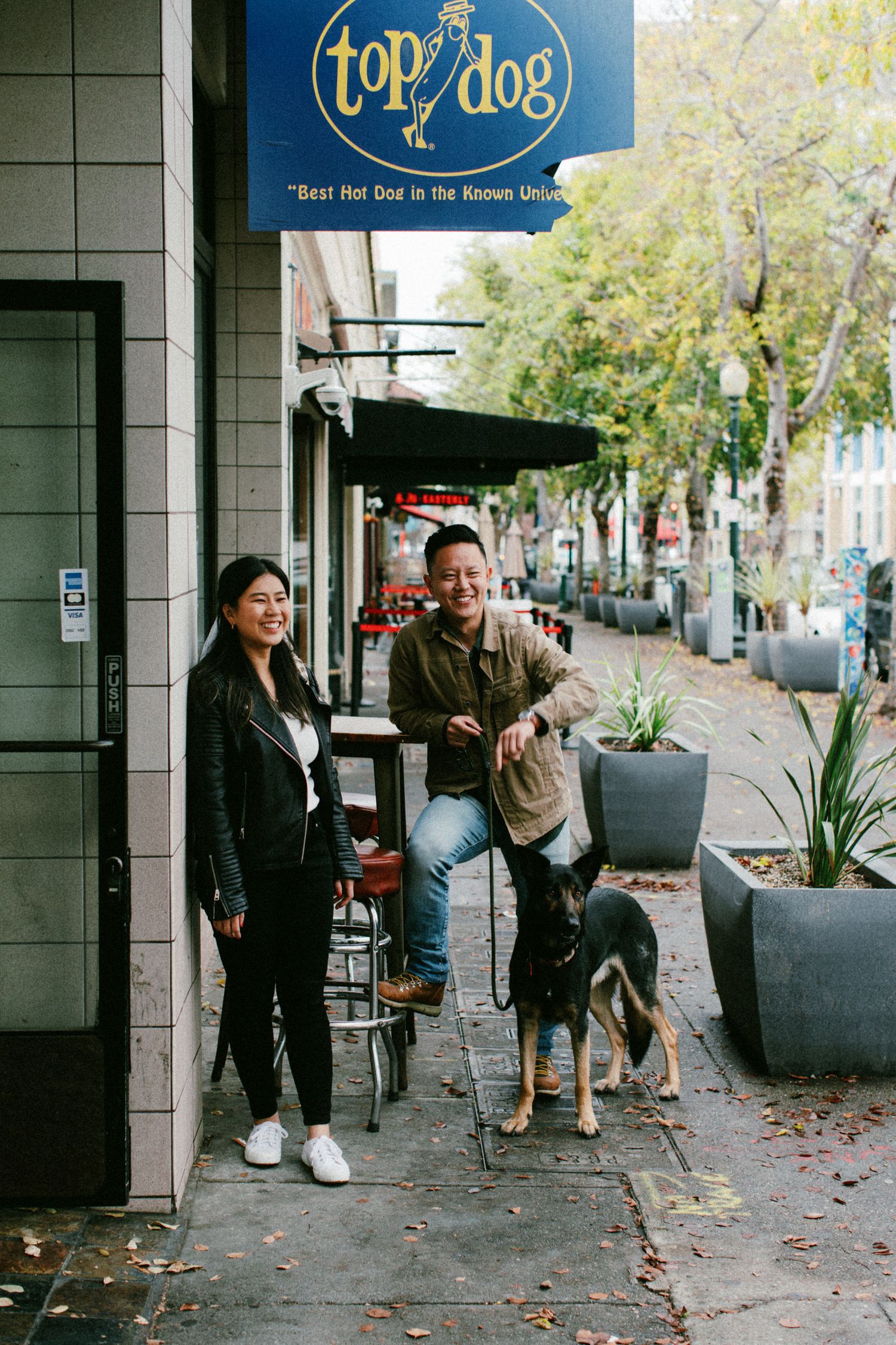 A couple stands with their dog outside a Top Dog restaurant on a tree-lined street.