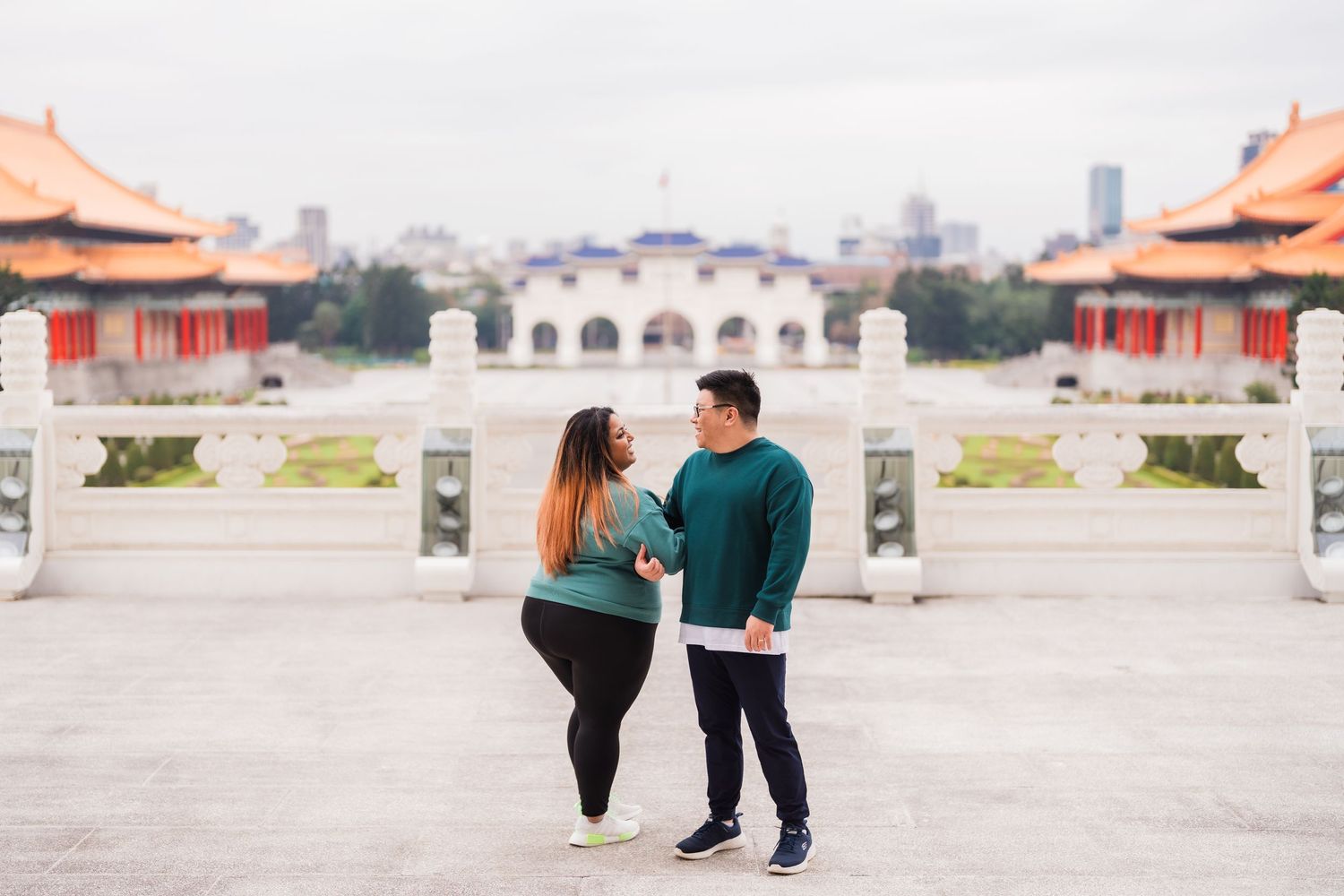 A couple poses together at a traditional Asian architectural site with city views in the background.