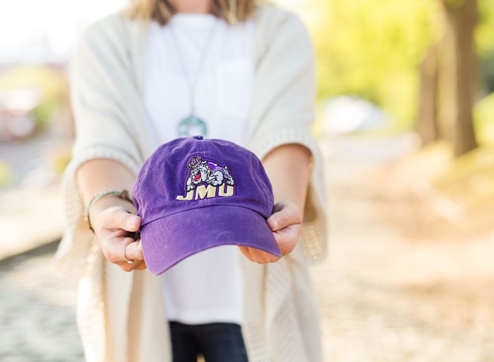 Hands holding a purple baseball cap with a team logo against a blurred outdoor background.