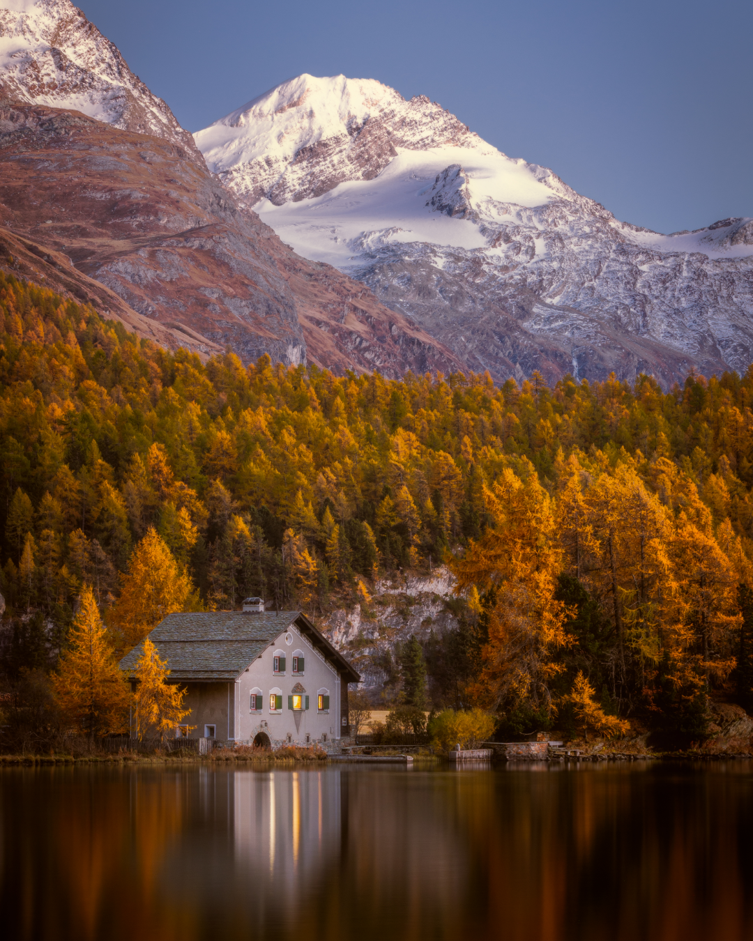 Alpine house reflecting in a calm lake with snow-covered mountain peaks and golden fall trees in background.