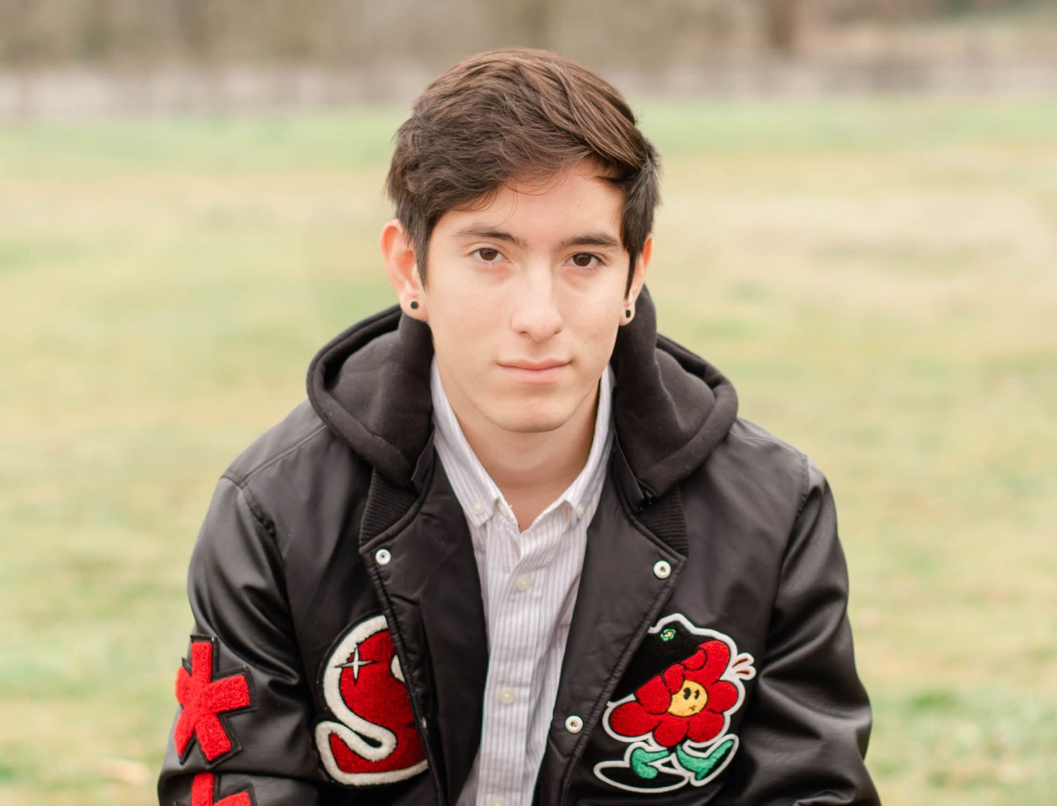 Person in black jacket with red patches poses outdoors in a casual setting.