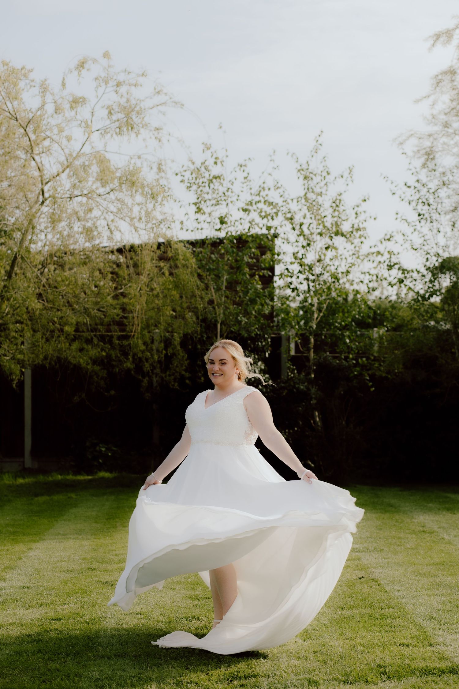 A bride in a flowing white dress twirls on a garden lawn with trees in the background.
