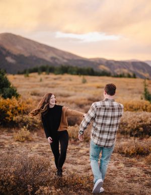 Two people running playfully through a mountain meadow at sunset.