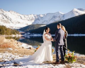 A winter wedding ceremony takes place beside a mountain lake with snow-capped peaks in the background.