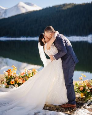 A wedding couple shares a romantic kiss by a mountain lake with orange floral decorations.