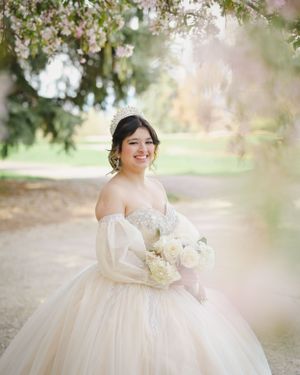 Bride in a flowing off-shoulder ballgown holds white bouquet under blossoming trees in outdoor portrait.