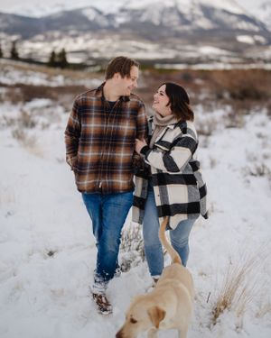 A couple walks with their dog through a snowy mountain landscape wearing plaid and buffalo check jackets.