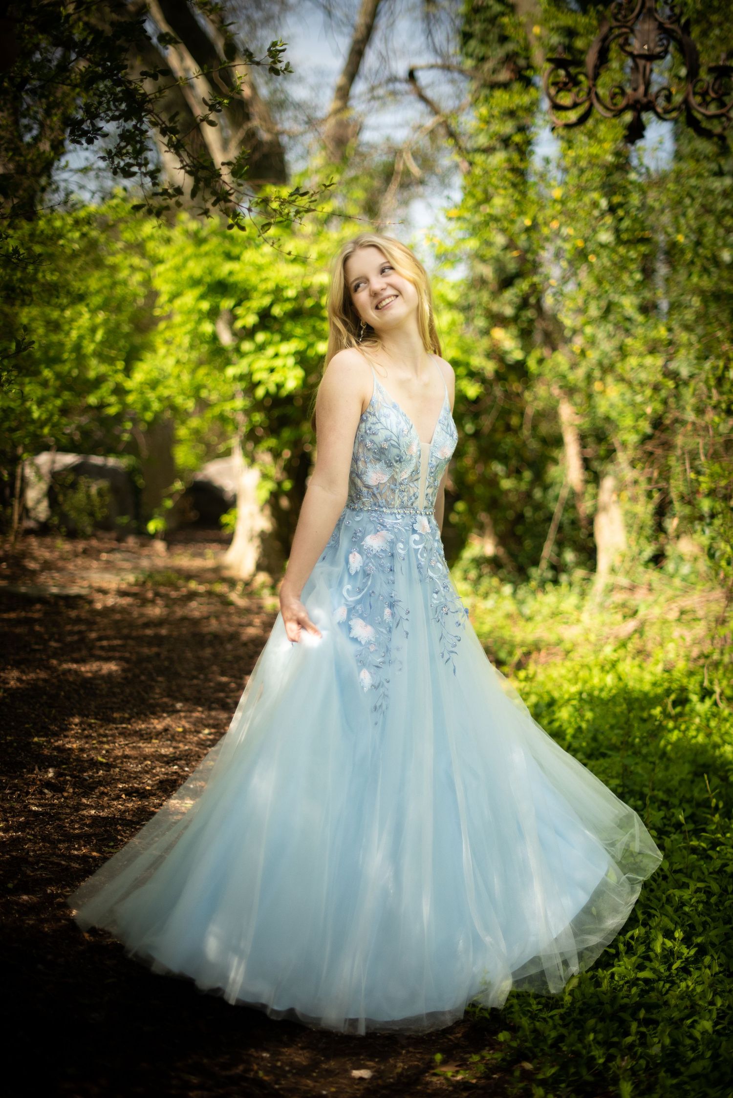 Person twirls in flowing light blue formal gown outdoors among spring foliage.