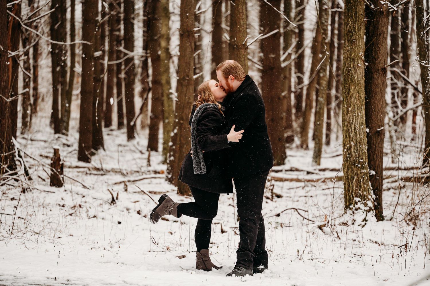 Couple sharing romantic kiss in snowy winter forest during engagement session.