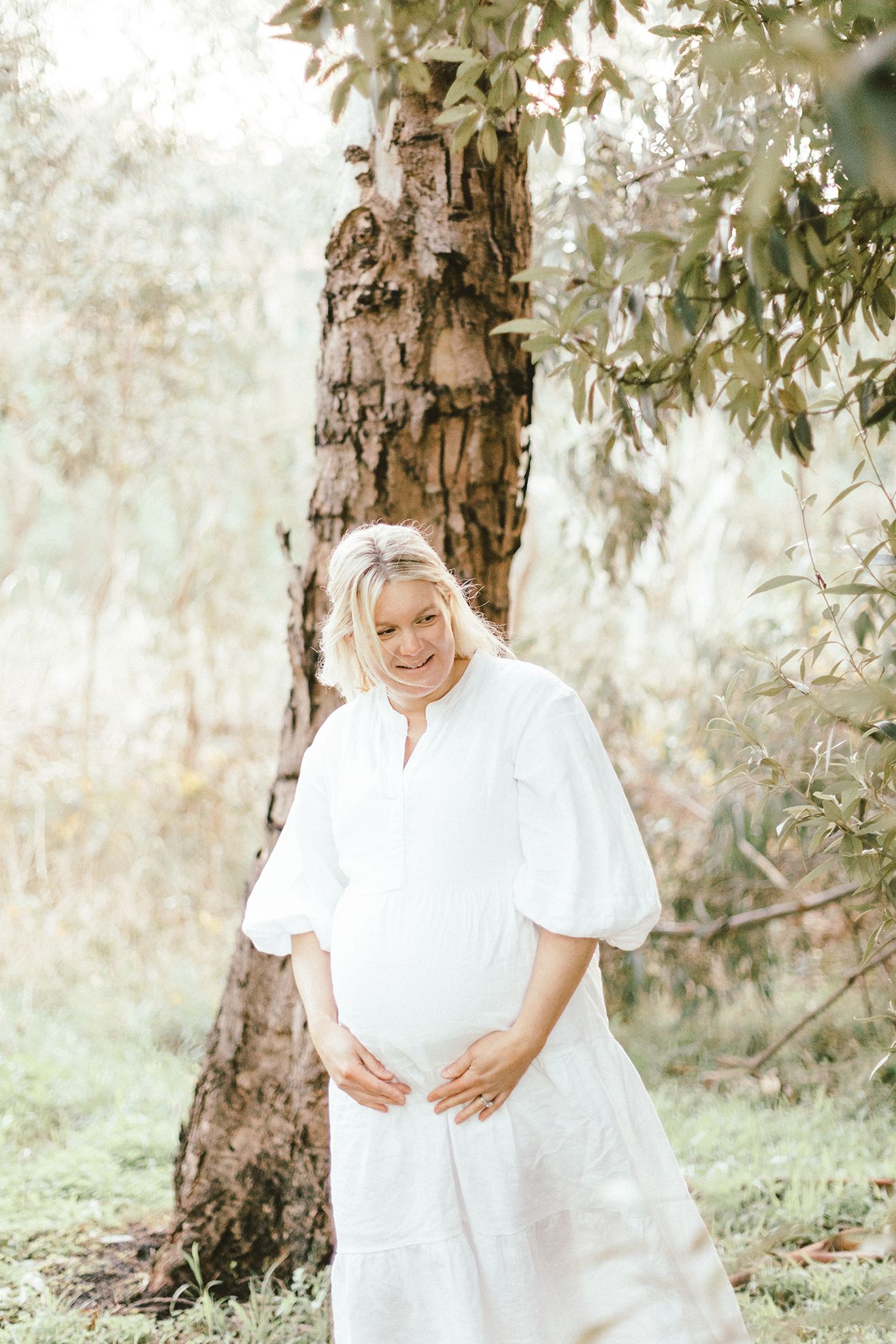 Expectant woman in flowing white dress standing by a tree trunk in a dreamy outdoor setting.