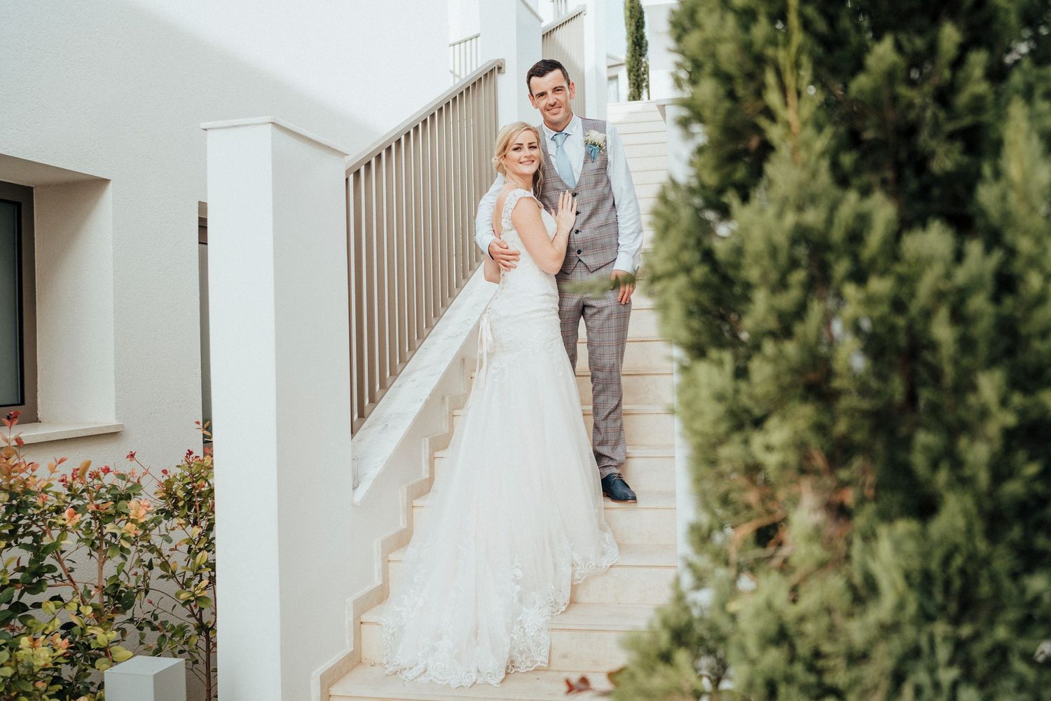 Newlyweds pose on white outdoor staircase surrounded by greenery and modern architecture.