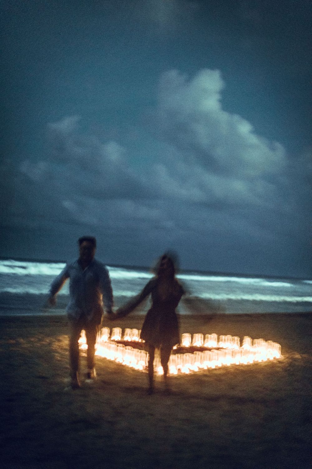 Couple standing by heart-shaped candle arrangement on beach at dusk.