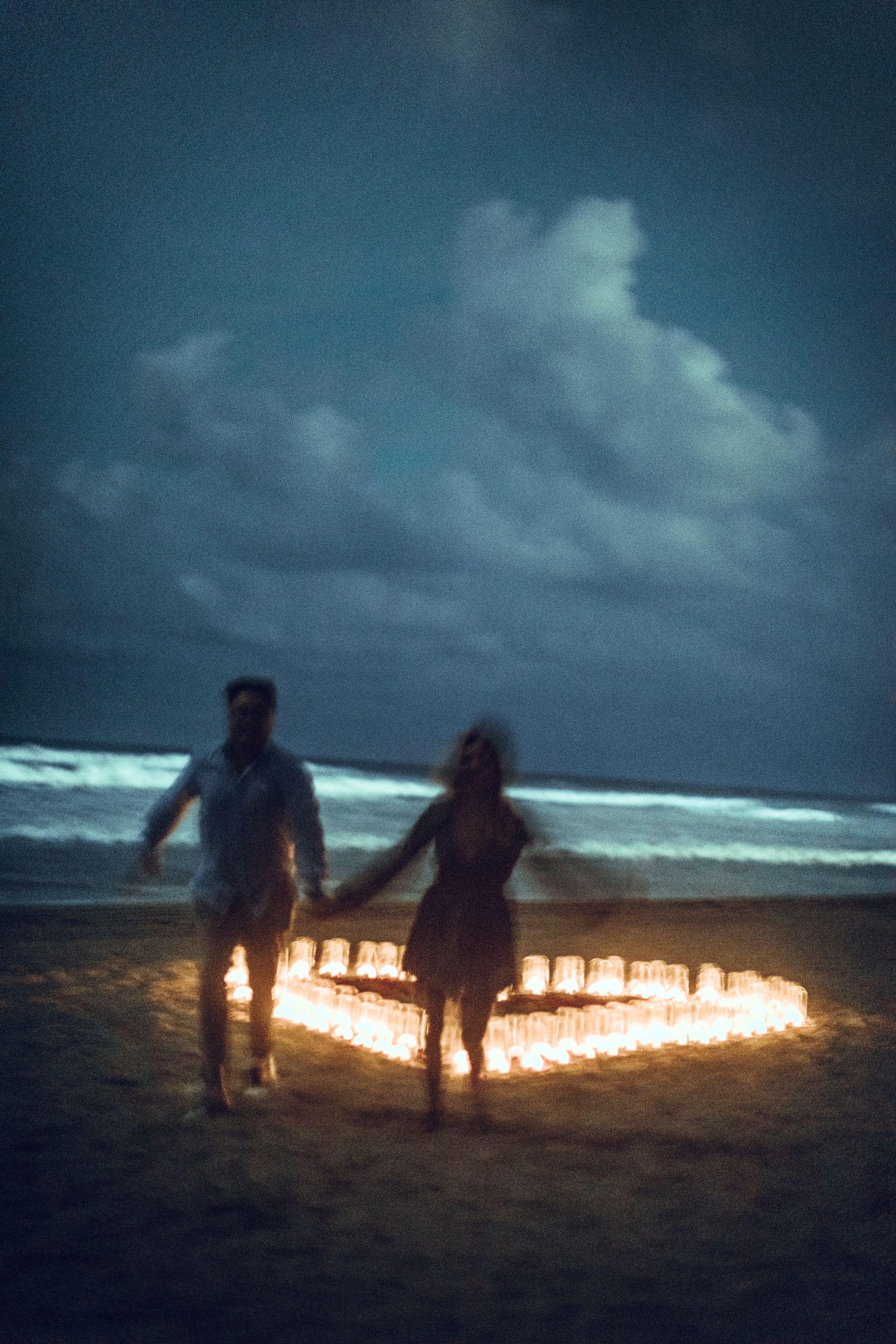Couple standing by heart-shaped candle arrangement on beach at dusk.