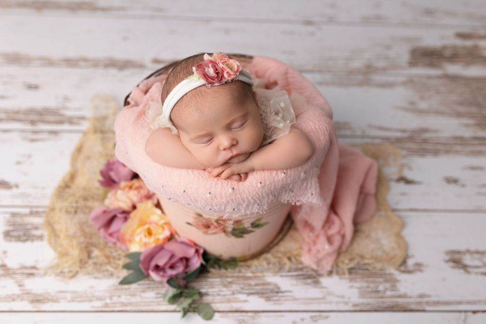 Sleeping newborn in pink bowl surrounded by flowers on rustic wooden backdrop during photo session.