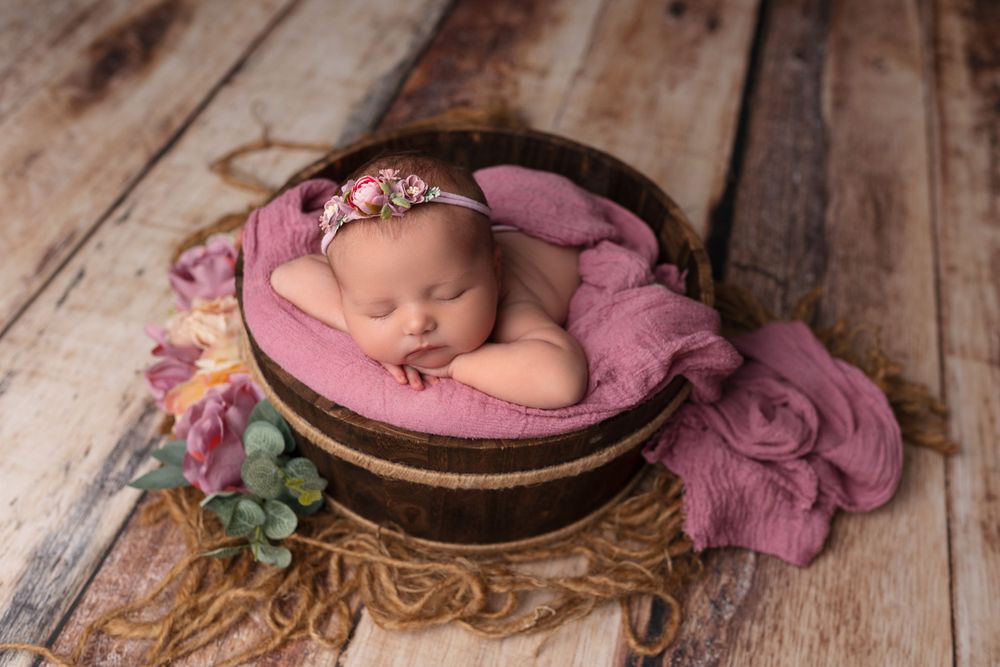 Newborn sleeping in pink cushioned bowl with floral decorations on wooden surface during photo shoot.