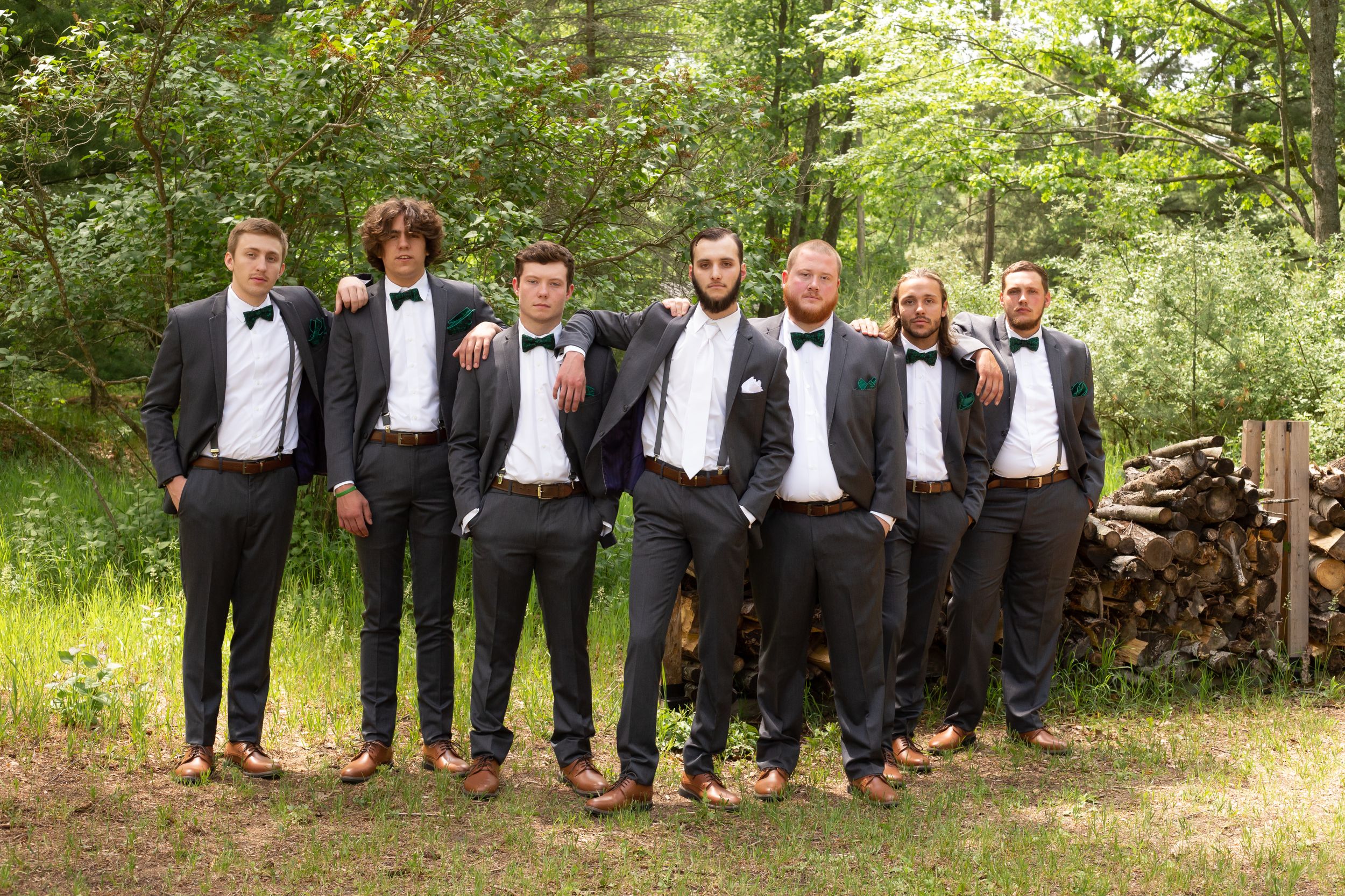 Groomsmen in black tuxedos and bowties pose together outdoors against a natural backdrop.