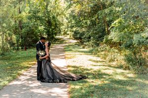 Couple in formal attire embraces on a sunlit garden path surrounded by greenery.