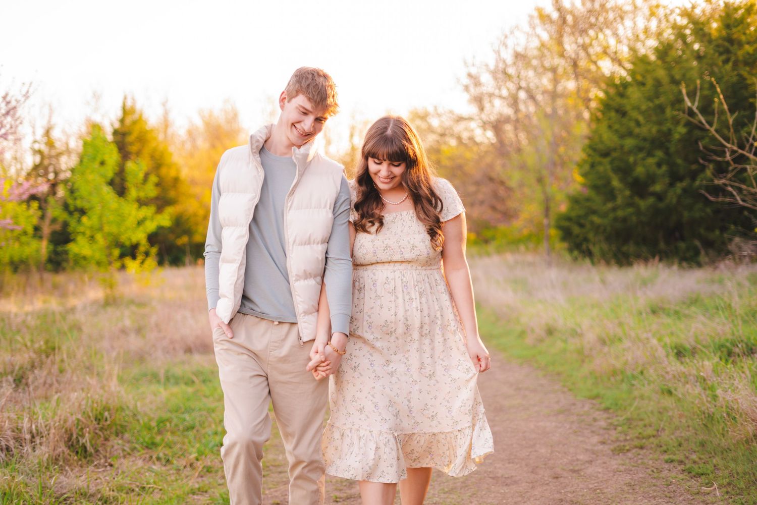 Happy couple walking hand in hand down a dirt path through golden meadow at sunset.