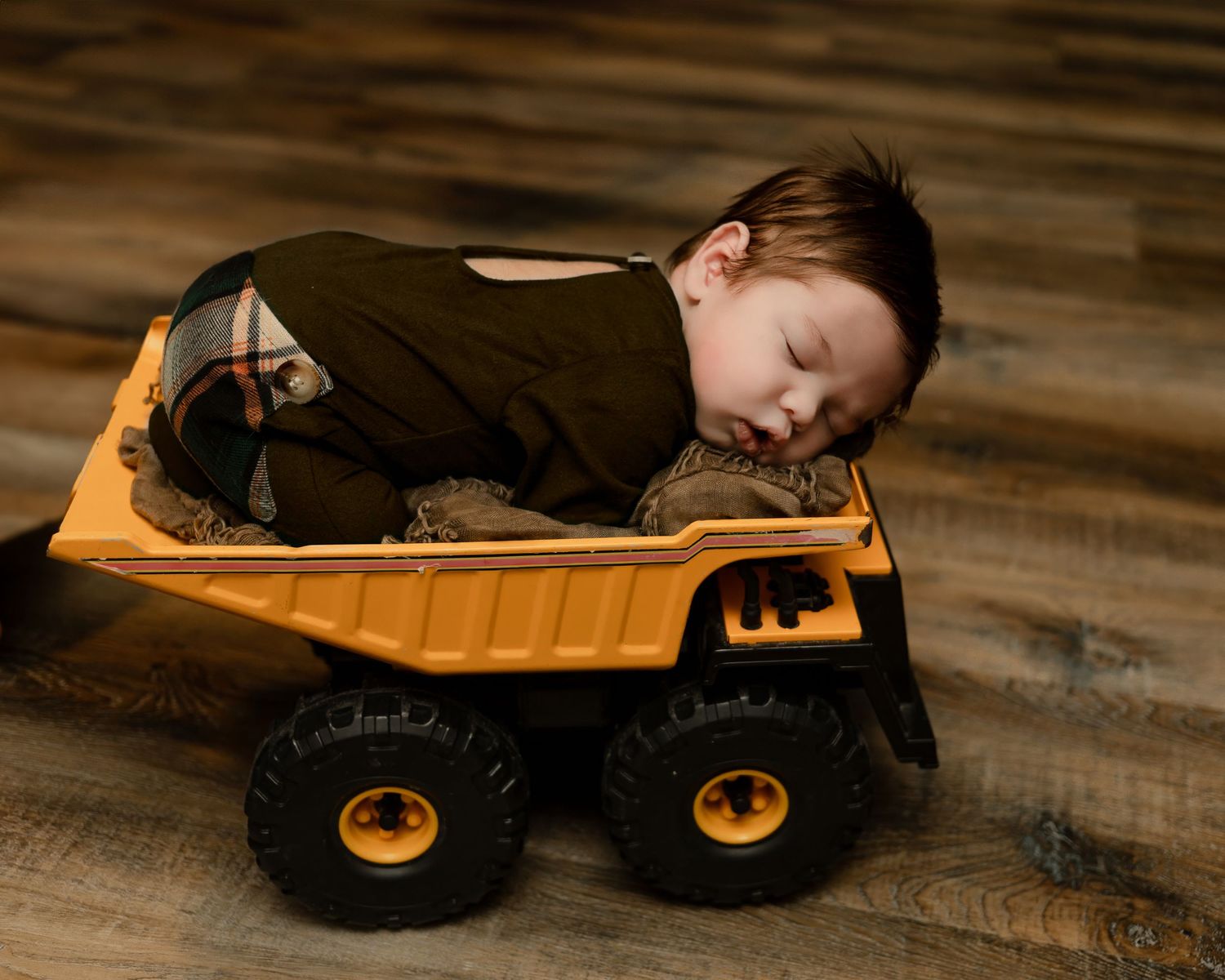 A sleeping child positioned in a yellow toy dump truck on wooden floor.