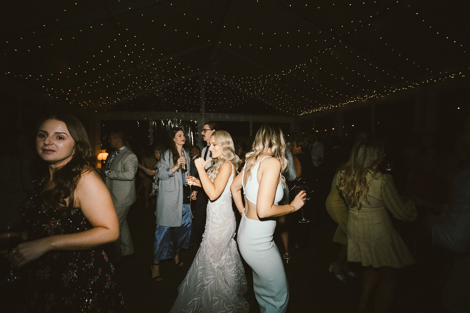 Wedding party dancing and celebrating on a dimly lit dance floor with string lights overhead.