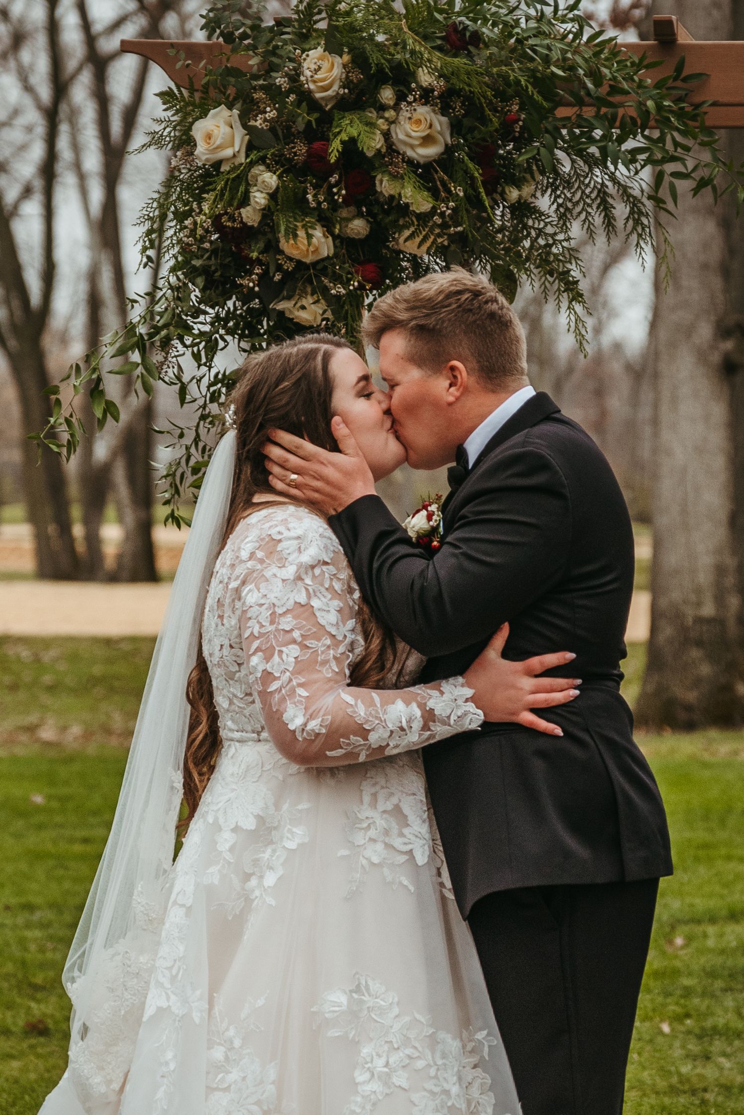 Wedding couple shares intimate kiss under floral arch in outdoor garden setting.