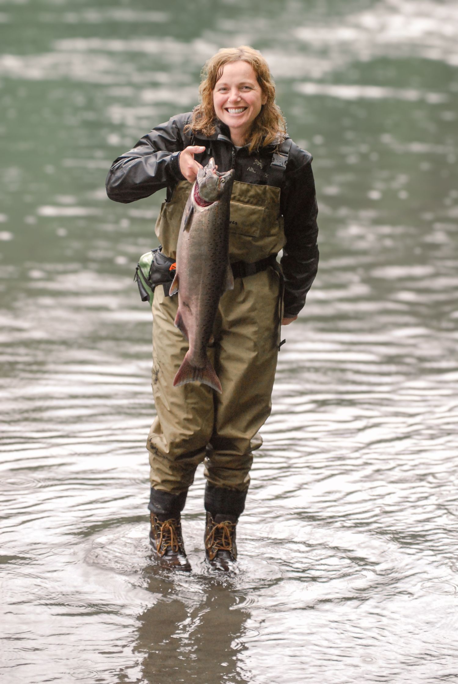 Person in waders stands in river holding a freshly caught fish while fly fishing.
