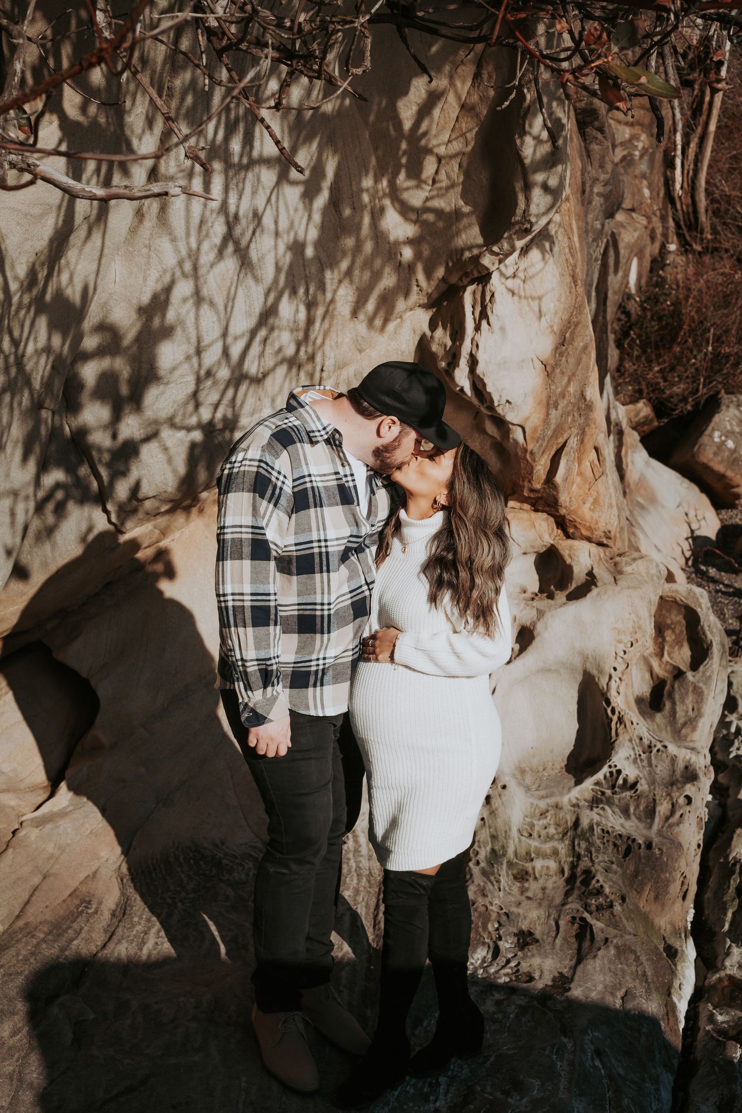Couple kissing and embracing beside textured sandstone cliffs, he in a plaid shirt and cap, she in a white sweater dress.