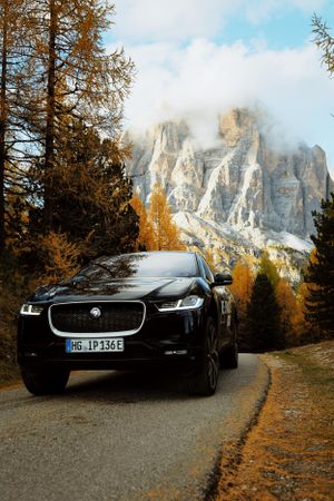 A sleek black Porsche sports car parked on a mountain road with autumn trees and dramatic Dolomite peaks in the background.
