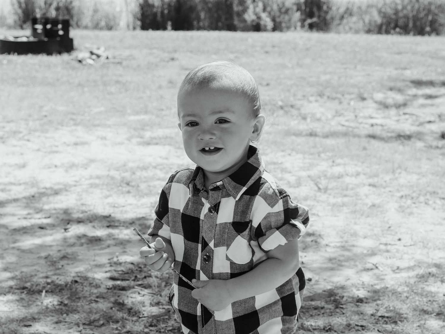 Cute smiling toddler boy in a plaid shirt standing in a sunny park.